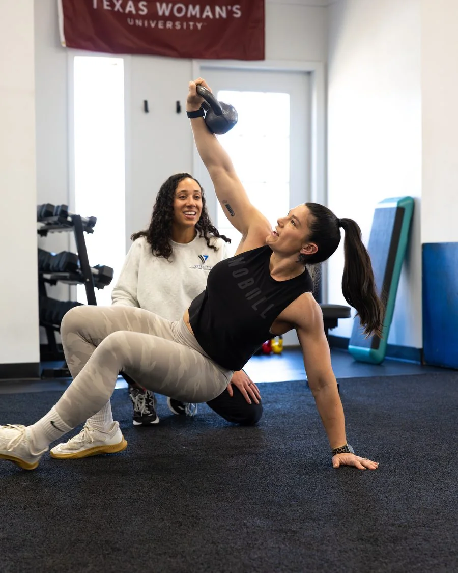 An athlete pressing a kettlebell overhead in a side plank with a Vitality coach
